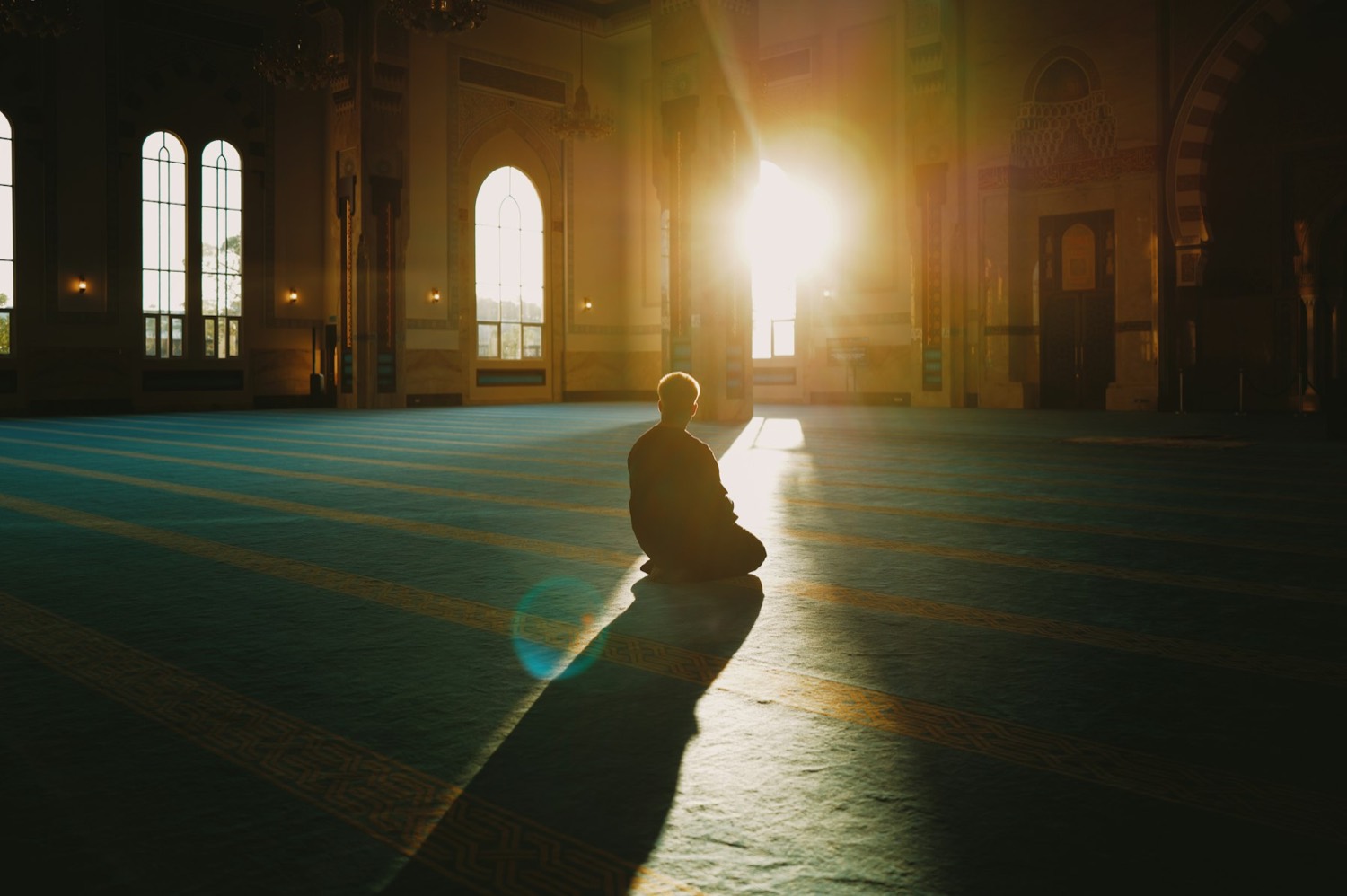 A man sitting alone in a large prayer hall as low sun pours through the doorway