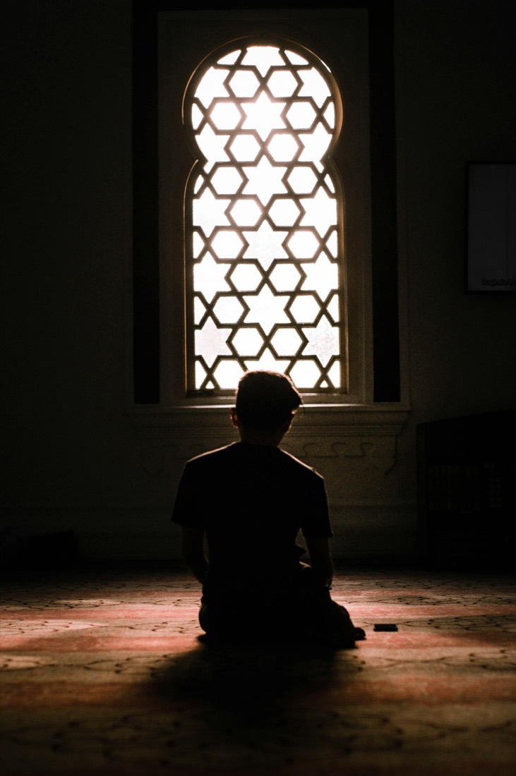 The silhouette of a person sitting in prayer before an arched lattice window