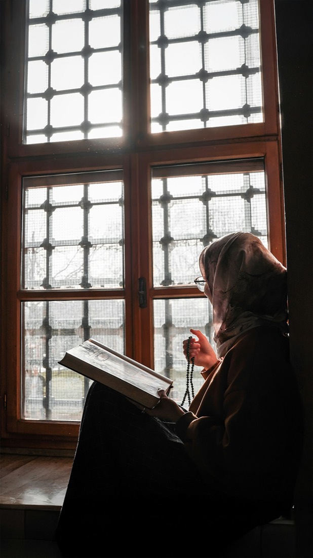 A woman in hijab reading by a latticed window, prayer beads in hand